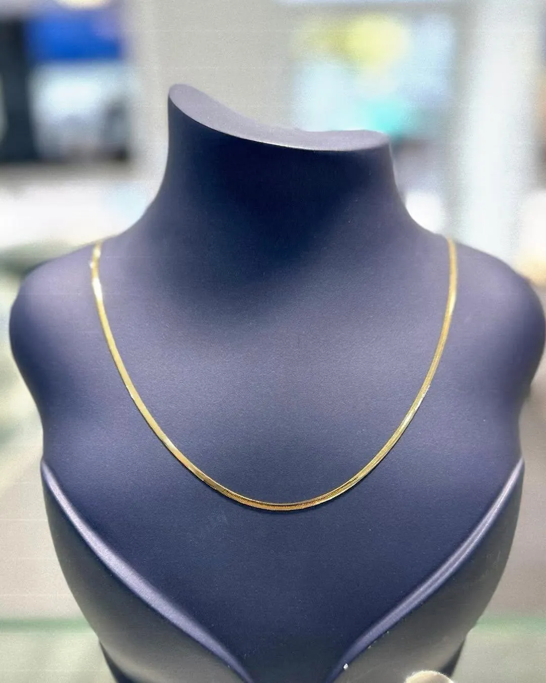 Schlanke Goldkette auf dunkelblauer Samtbüste, im Hintergrund unscharf der Laden