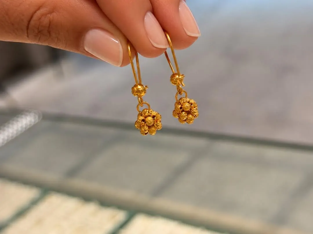 Zwei goldene Ohrhänger mit Kugelblüten zwischen zwei Fingern, darunter unscharf die Tabletts einer Ladenvitrine