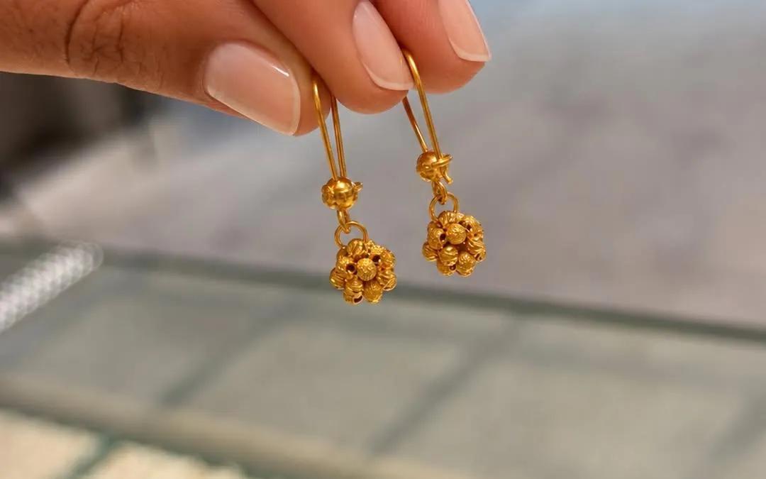 Zwei goldene Ohrhänger mit Kugelblüten zwischen zwei Fingern, darunter unscharf die Tabletts einer Ladenvitrine (mobiler Ausschnitt)